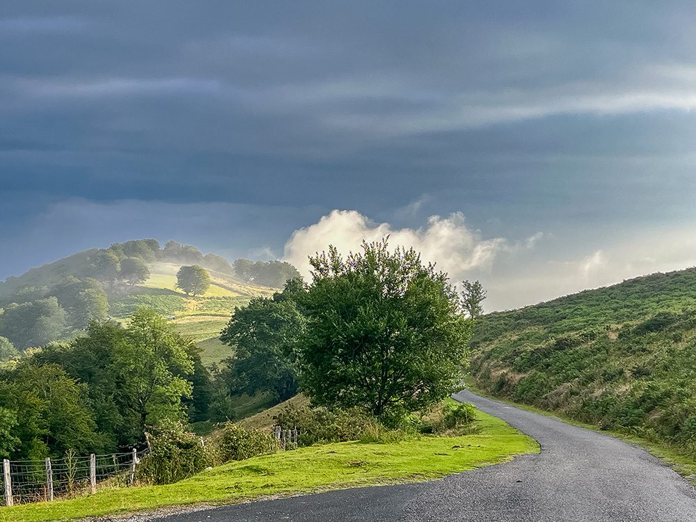 Mountain road through the Pyrenees on the Camino Francés between Borda and Roncesvalles, with sunlight breaking through dramatic clouds.