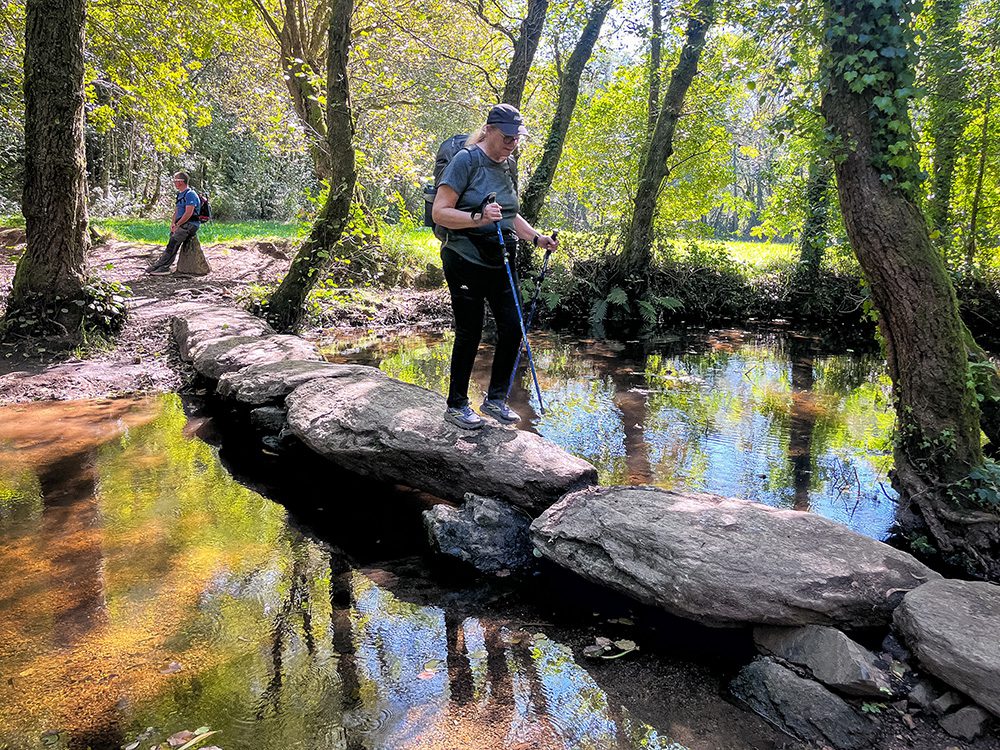 Pilgrim crossing stepping stones over a woodland stream on the Camino Francés in Galicia