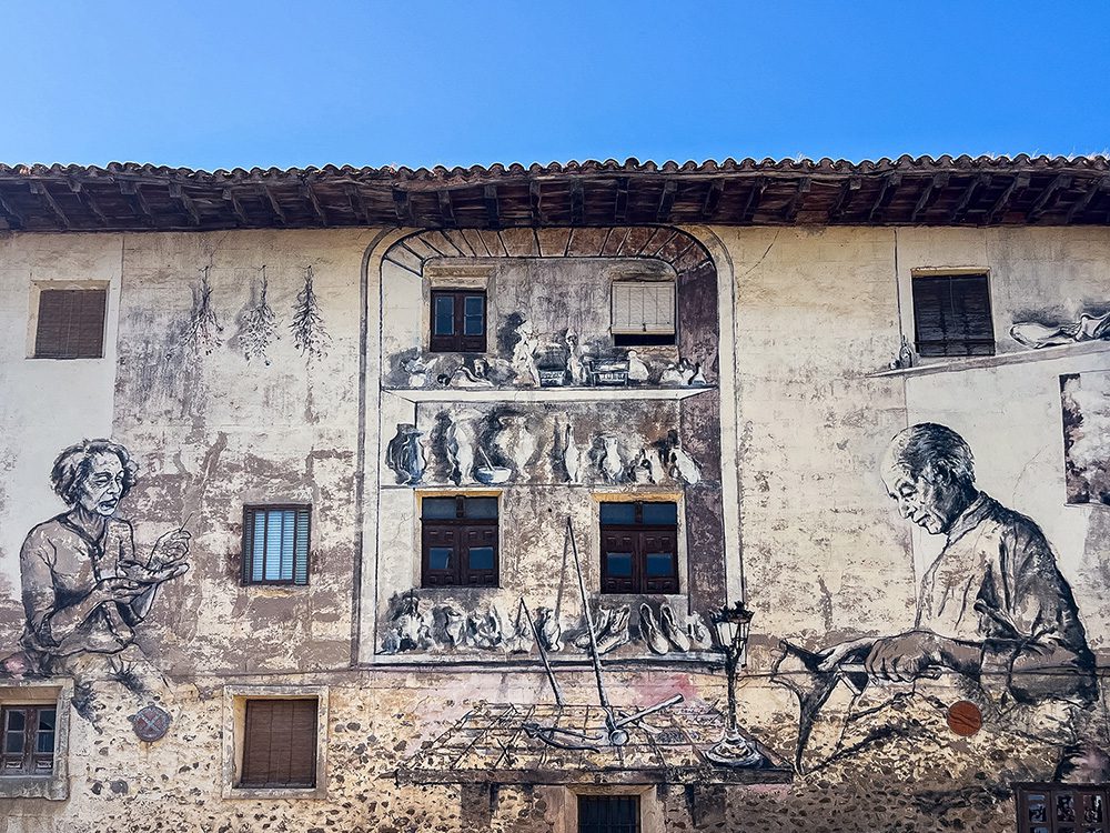 Large street art mural depicting traditional craftspeople on a building in Belorado