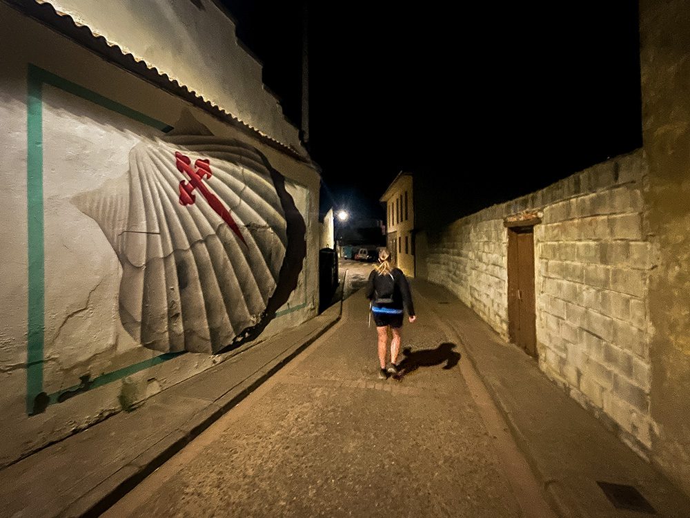 Pilgrim walking past a giant scallop shell mural while leaving Sahagún before dawn on the Camino Francés